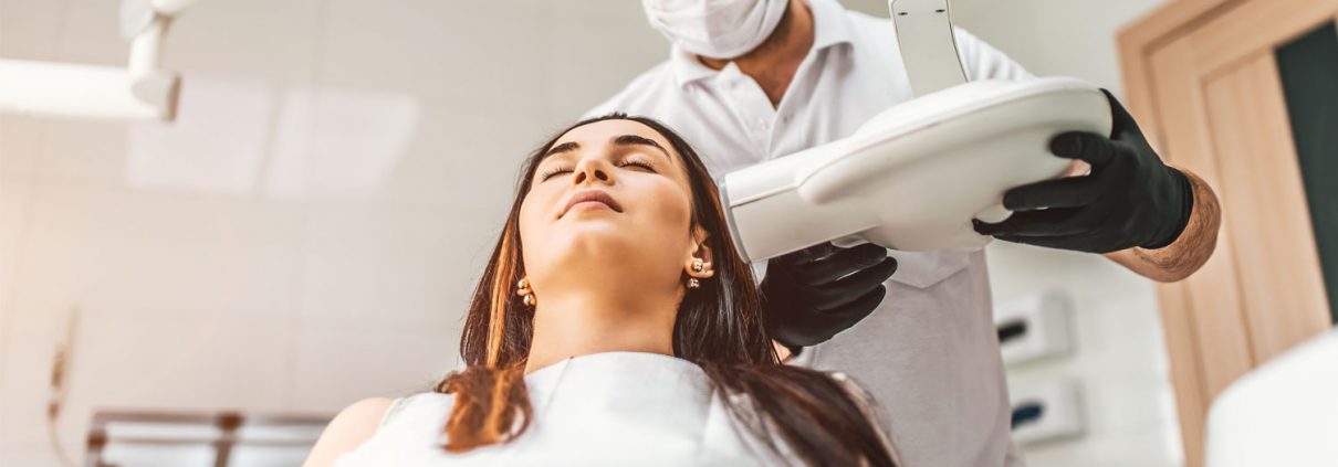 A dental technician prepares to take an X-ray of a woman sitting in a dental chair.