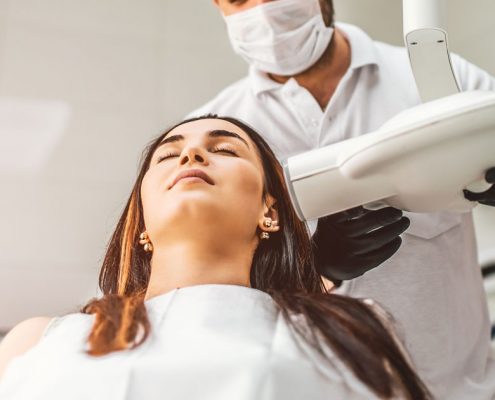A dental technician prepares to take an X-ray of a woman sitting in a dental chair.