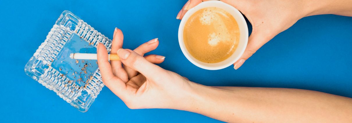 A person holds a cup of coffee in one hand and a lit cigarette over an ashtray in the other, against a blue background.