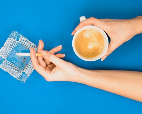 A person holds a cup of coffee in one hand and a lit cigarette over an ashtray in the other, against a blue background.