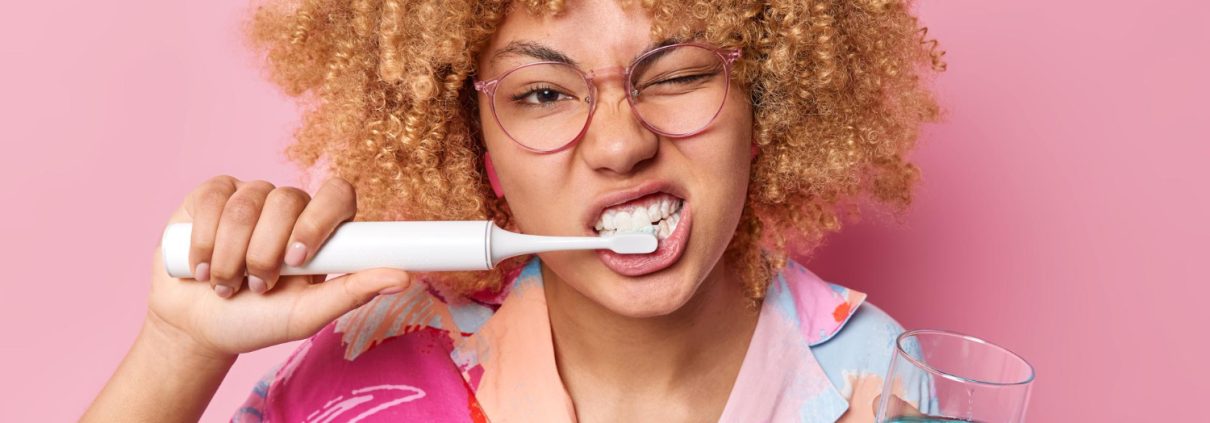 Person with curly hair and glasses brushes teeth with an electric toothbrush while holding a glass of blue mouthwash, against a pink background.