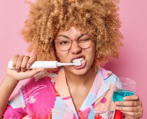 Person with curly hair and glasses brushes teeth with an electric toothbrush while holding a glass of blue mouthwash, against a pink background.
