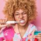 Person with curly hair and glasses brushes teeth with an electric toothbrush while holding a glass of blue mouthwash, against a pink background.