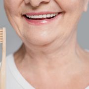 Close-up of a smiling older woman holding a wooden toothbrush, with focus on her mouth and teeth, against a plain gray background.
