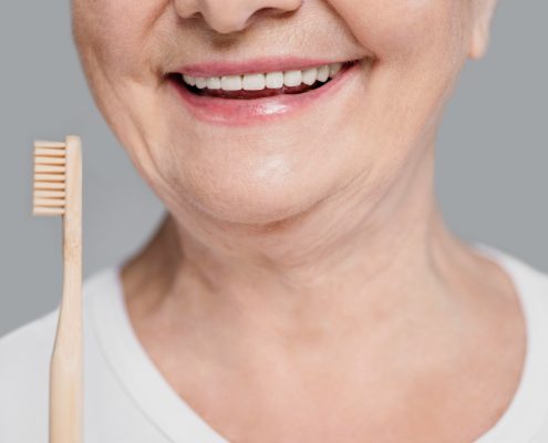 Close-up of a smiling older woman holding a wooden toothbrush, with focus on her mouth and teeth, against a plain gray background.