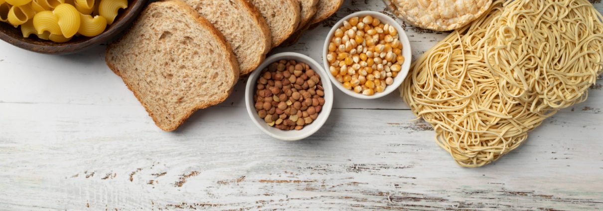 Assorted carbohydrates including pasta, sliced bread, rice cakes, noodles, grains, and legumes arranged on a white wooden surface.
