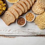 Assorted carbohydrates including pasta, sliced bread, rice cakes, noodles, grains, and legumes arranged on a white wooden surface.