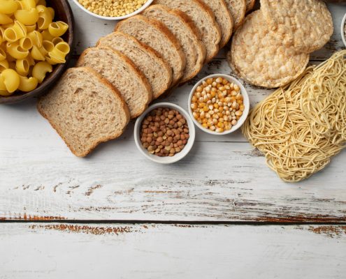 Assorted carbohydrates including pasta, sliced bread, rice cakes, noodles, grains, and legumes arranged on a white wooden surface.