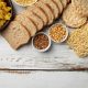 Assorted carbohydrates including pasta, sliced bread, rice cakes, noodles, grains, and legumes arranged on a white wooden surface.