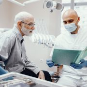 A dentist wearing gloves and a mask discusses a patients file with an older man in a dental office.