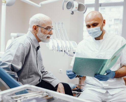 A dentist wearing gloves and a mask discusses a patients file with an older man in a dental office.