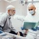 A dentist wearing gloves and a mask discusses a patients file with an older man in a dental office.