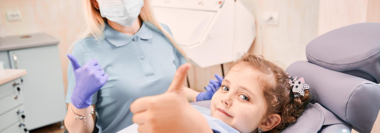 A female dentist wearing a mask and gloves sits next to a young girl in a dental chair; both are giving thumbs up to the camera.