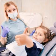 A female dentist wearing a mask and gloves sits next to a young girl in a dental chair; both are giving thumbs up to the camera.