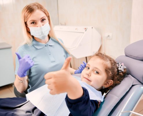 A female dentist wearing a mask and gloves sits next to a young girl in a dental chair; both are giving thumbs up to the camera.