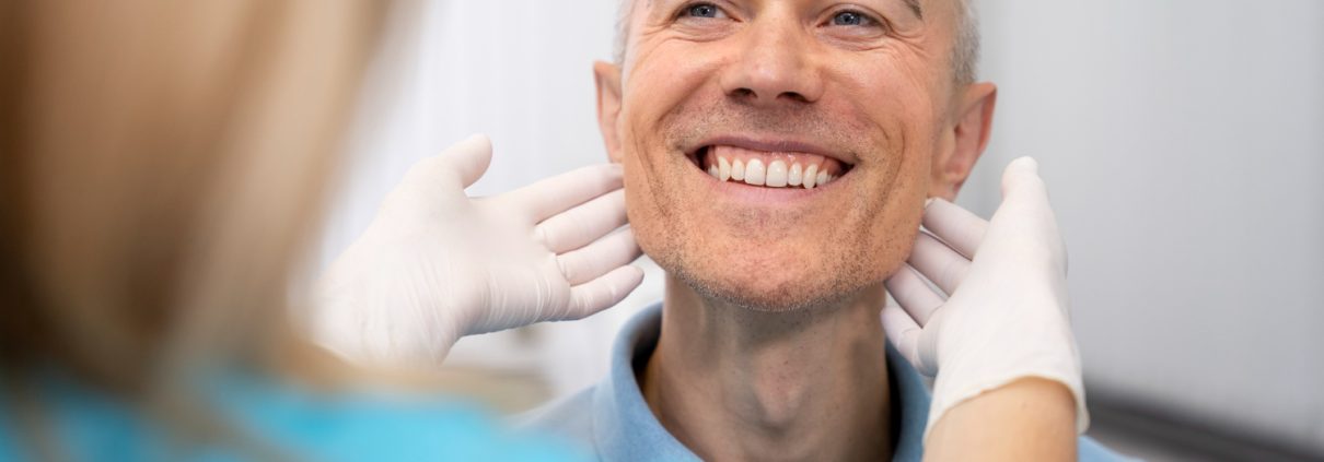 A healthcare professional wearing gloves examines a smiling man’s jawline and neck during a medical checkup.