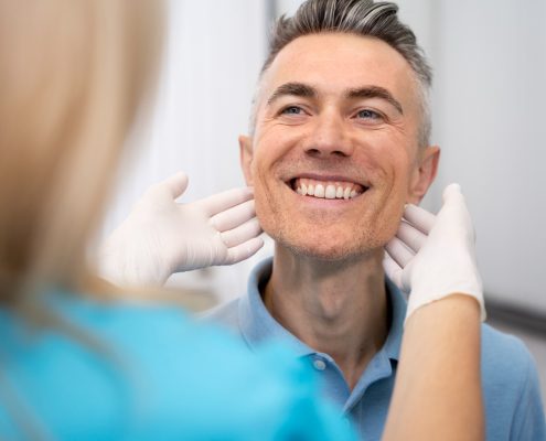 A healthcare professional wearing gloves examines a smiling man’s jawline and neck during a medical checkup.
