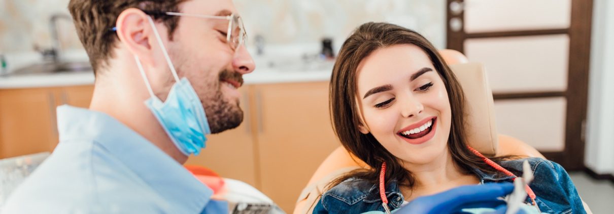 A dentist wearing gloves and a mask shows a dental tool to a smiling female patient seated in a dental chair.