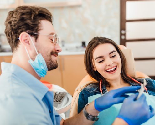A dentist wearing gloves and a mask shows a dental tool to a smiling female patient seated in a dental chair.