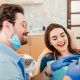 A dentist wearing gloves and a mask shows a dental tool to a smiling female patient seated in a dental chair.