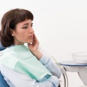 A woman sits in a dentist chair, touching her cheek with a concerned expression, wearing a dental bib in a clinic setting.