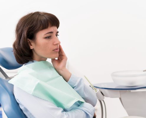 A woman sits in a dentist chair, touching her cheek with a concerned expression, wearing a dental bib in a clinic setting.