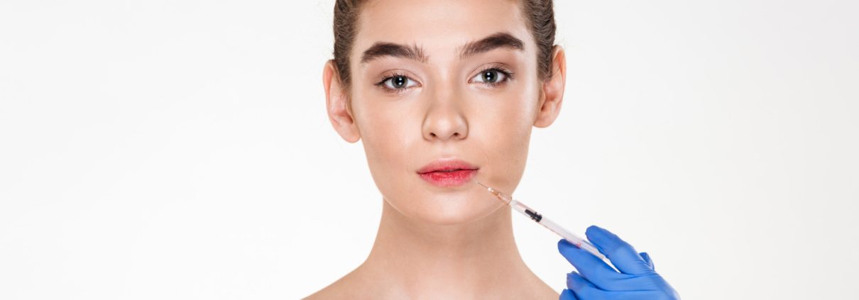 A woman with bare shoulders faces forward while a gloved hand holds a syringe near her lips, suggesting a cosmetic injection procedure.