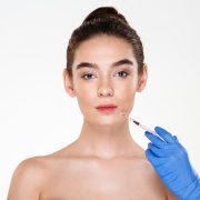 A woman with bare shoulders faces forward while a gloved hand holds a syringe near her lips, suggesting a cosmetic injection procedure.