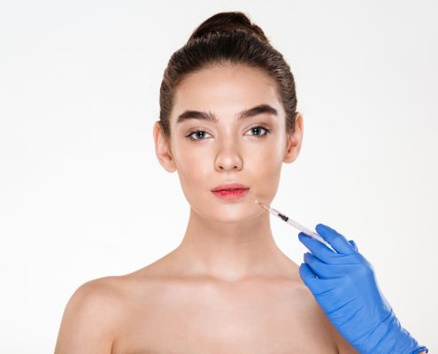 A woman with bare shoulders faces forward while a gloved hand holds a syringe near her lips, suggesting a cosmetic injection procedure.